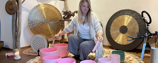 Junge Frau sitzt im Meditationsraum umgeben von Klangschalen, Gongs und Ornamenten.