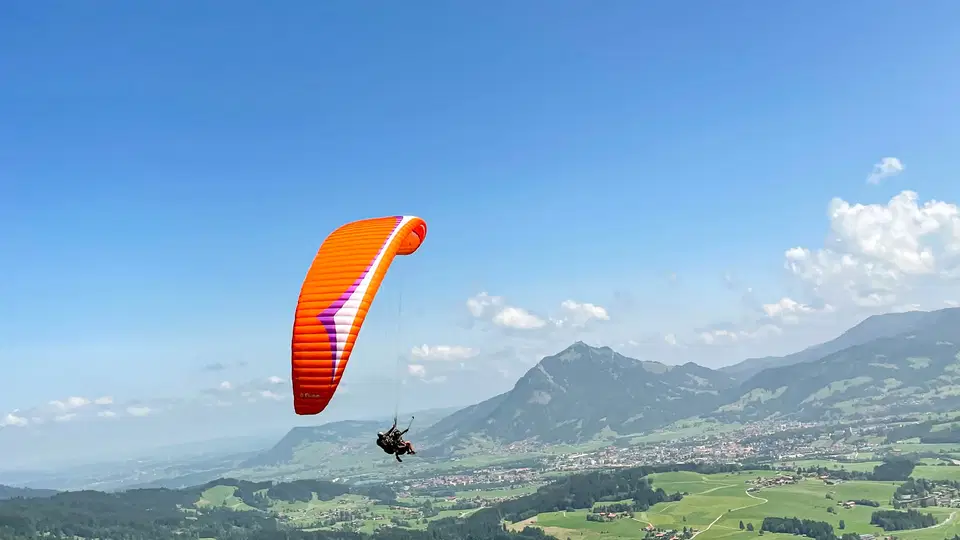 Orangefarbener Gleitschirm über grünen Wiesen und dem Tal.