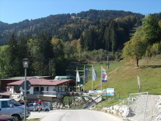 Kleine Berghütte am Hang mit Fahnen vor Waldlandschaft, davor Parkplatz