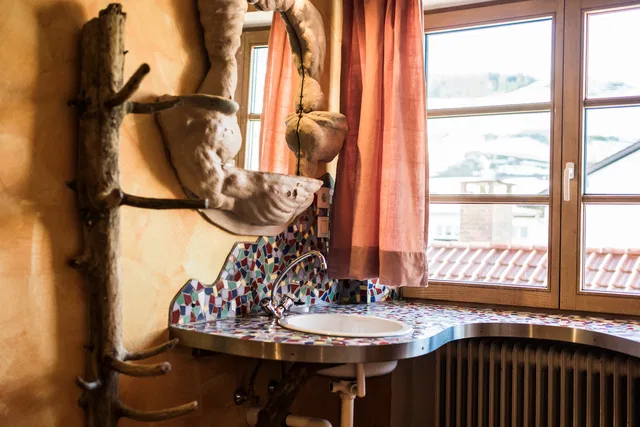 Waschtisch mit bunten Mosaikfliesen, Fenster mit Vorhängen und Heizkörper darunter, Wanddeko aus Äst