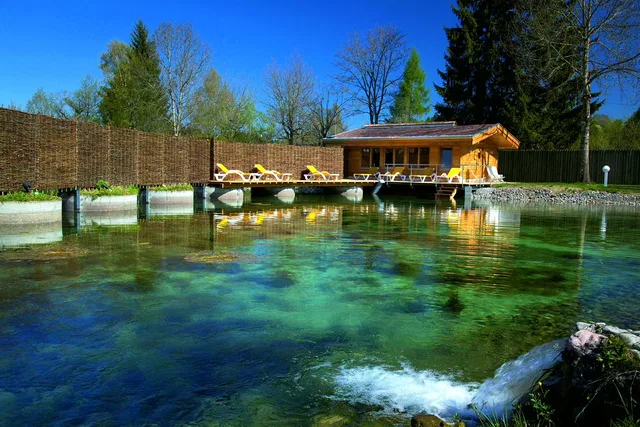 Holzhaus am Teich mit Liegen auf dem Steg, umgeben von Bäumen.