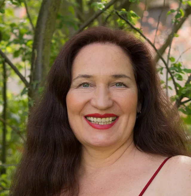 Porträt einer Frau mit langen braunen Haaren, lächelnd vor grünem Garten.