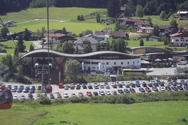 Gebäudekomplex Steinplatte mit welliger Dachlinie, Parkplatz voller Autos vor grüner Berglandschaft.