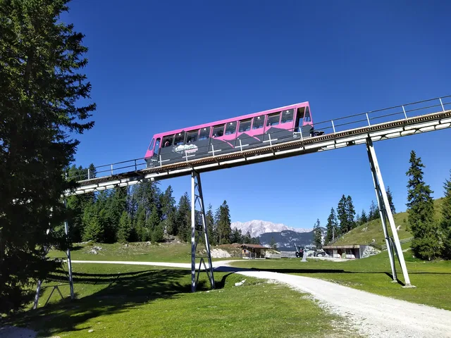 Pinkfarbener Zug fährt auf einer Brückenbrücke über grüne Wiesen, Berge im Hintergrund.