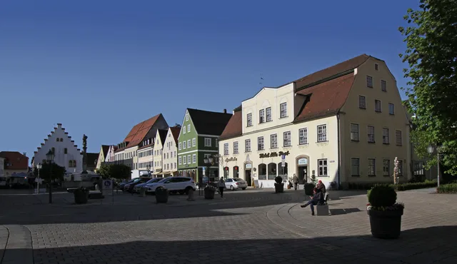 Bunte Häuserzeile am Marktplatz mit Cafés und Passanten unter klarem Himmel.