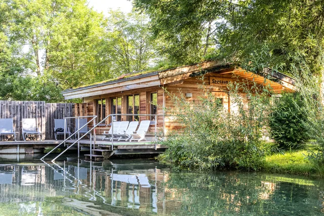 Holzhaus am See mit Terrasse, Liegen und Stufen ins Wasser, von Bäumen umgeben.