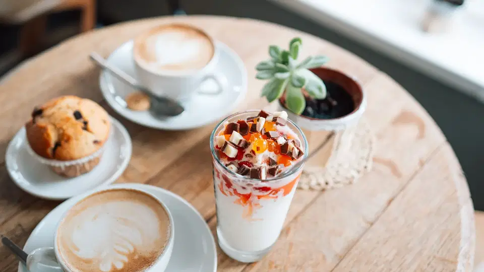 Runder Holztisch im Café mit Latte-Art, Cappuccino, Muffin, Joghurt-Getränk und Topfpflanze.