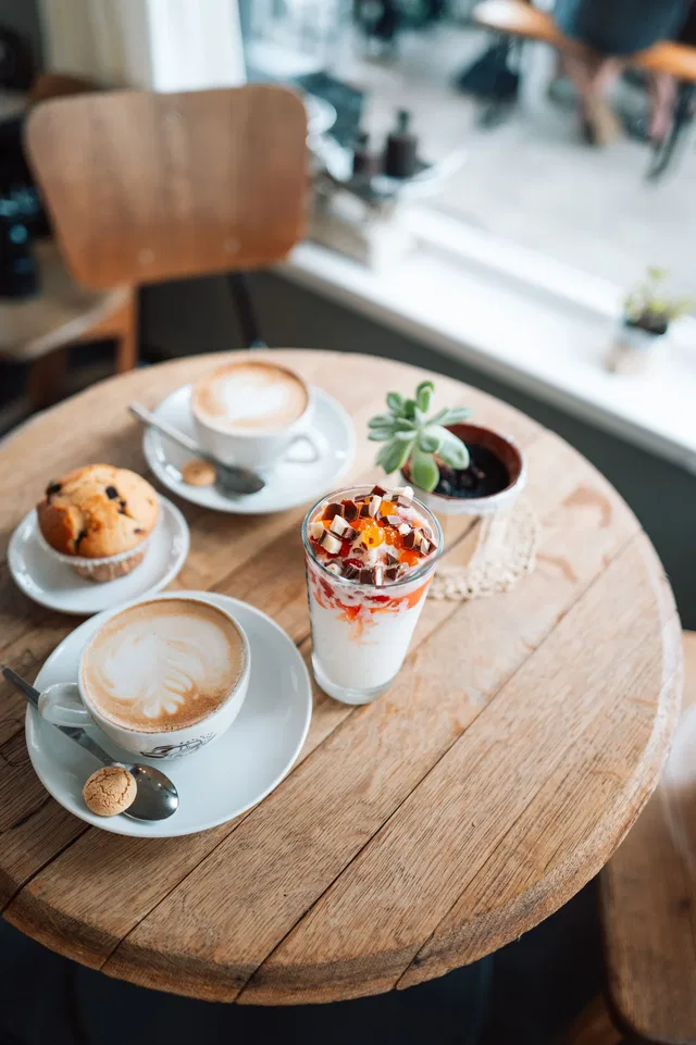 Runder Holztisch im Café mit Latte-Art, Cappuccino, Muffin, Joghurt-Getränk und Topfpflanze.