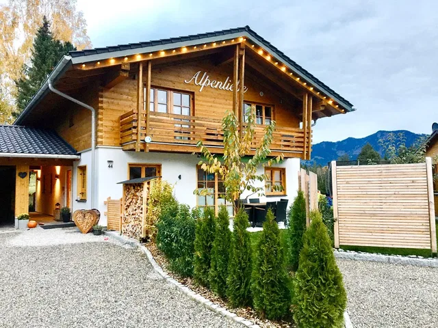 Holzhaus im alpinen Stil mit Balkon, davor Hecke aus Koniferen und gepflasterter Weg.