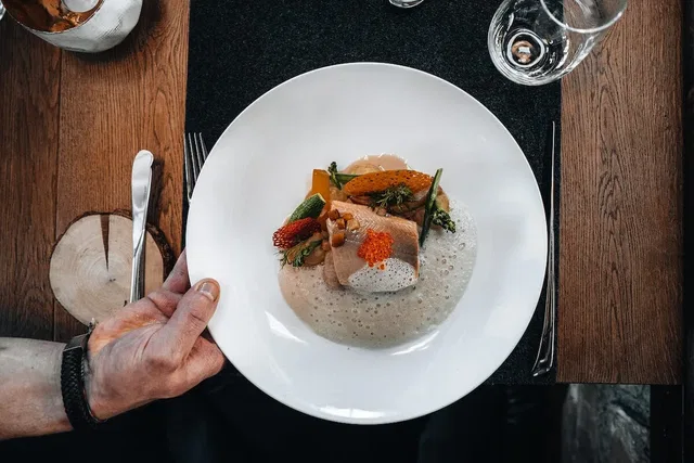 Hand hält einen weißen Teller mit Fisch, Gemüse und Schaum von oben