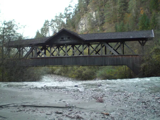 Überdachte Holzkbrücke mit Fachwerk über einem Fluss im Wald.