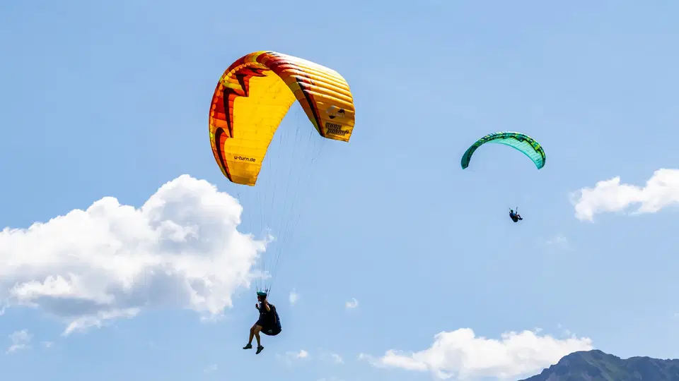 Zwei Gleitschirmflieger vor blauem Himmel und Bergsilhouette.