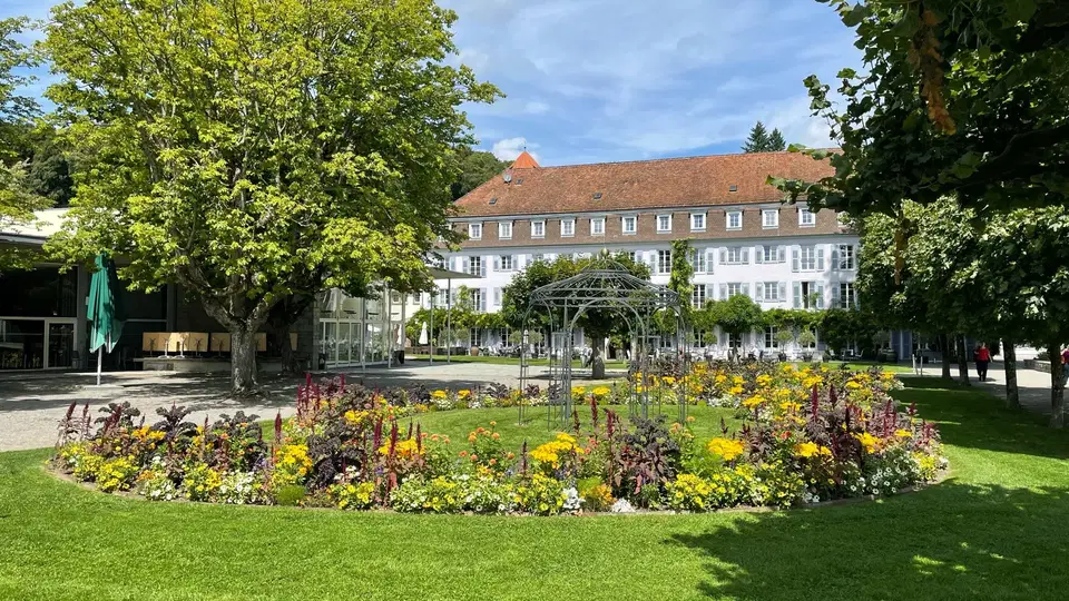 Gepflegter Innenhof mit Blumenbeeten, Bäumen und Metallpergola vor einem mehrstöckigen Gebäudekomple