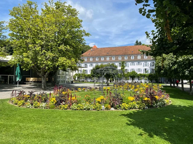 Gepflegter Innenhof mit Blumenbeeten, Bäumen und Metallpergola vor einem mehrstöckigen Gebäudekomple