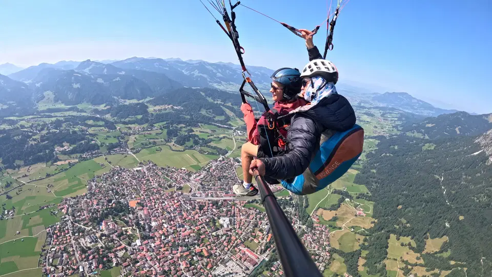 Gast in roter Jacke und Pilot beim Tandemflug über dem Ort im Tal.