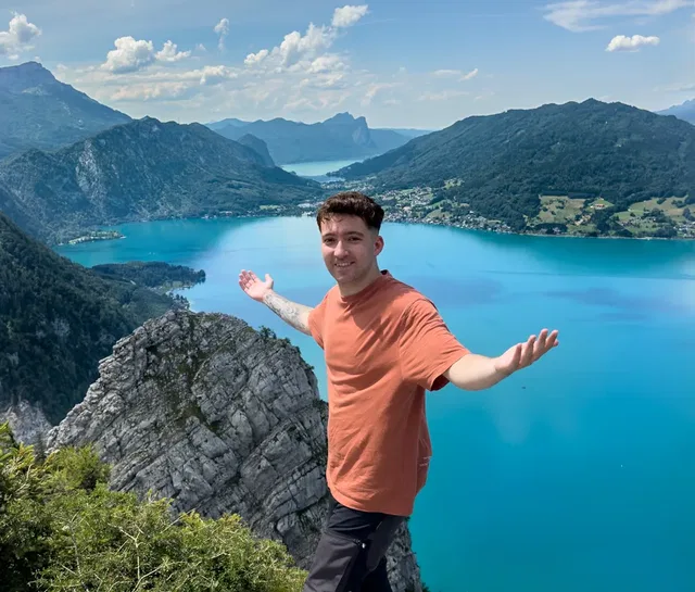 Mann mit orangefarbenem T-Shirt vor türkisblauem See und Berglandschaft