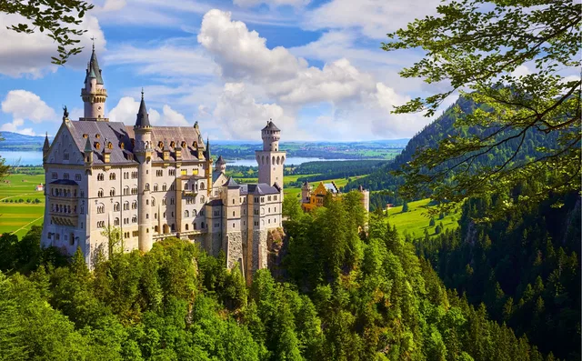 Schloss Neuschwanstein thront auf einem Waldhügel mit Bergen im Hintergrund.