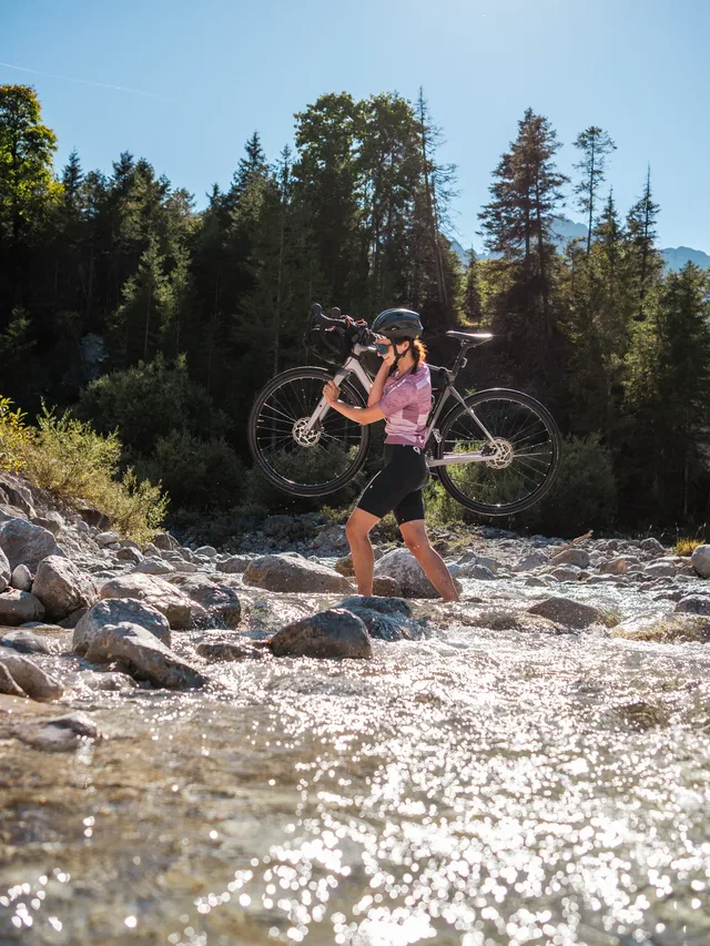 Person trägt Gravelbike über Kies am Flussufer mit Wald im Hintergrund.