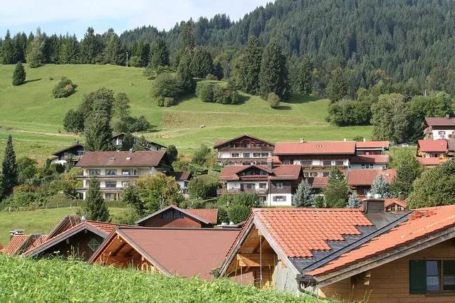 Hügeliges Alpendorf mit Holzhäusern und Rotziegeldächern vor Wald und Grünland.