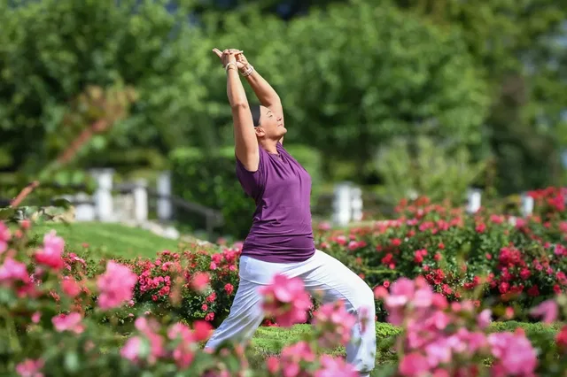 Frau in lila Oberteil und weißen Hosen übt Yoga in einem Rosengarten.