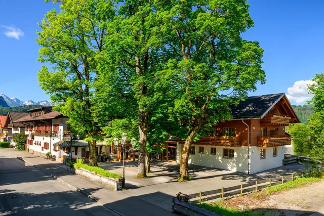 Häuser am Dorfplatz mit großen Laubbäumen und Berglandschaft im Hintergrund.