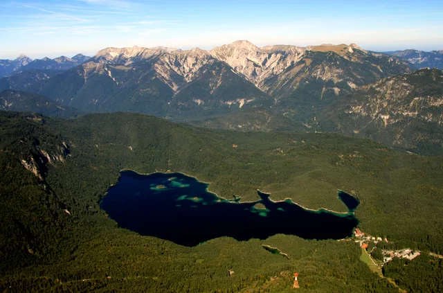 Luftaufnahme eines grünen Gebirgslands mit dunklem Bergsee im Tal und umliegenden Bergen.