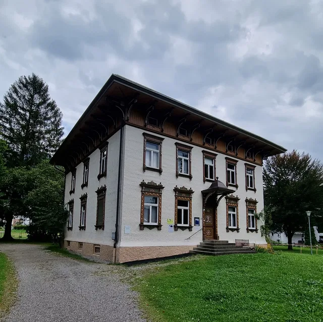 Historisches zweistöckiges Haus mit verzierten Fensterrahmen und Vordach vor Grünfläche.