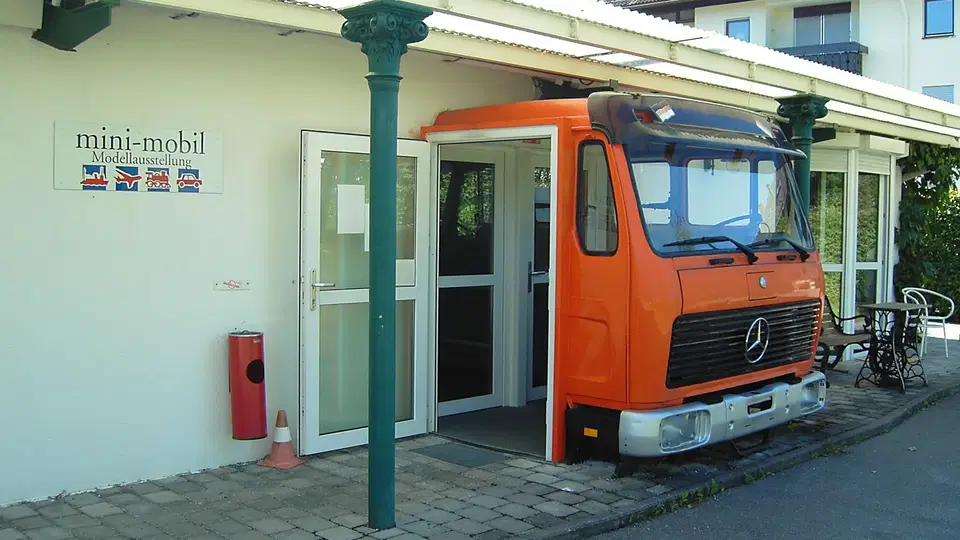 Orangener Mercedes-LKW vor offener Eingangstür eines Ausstellungsraums