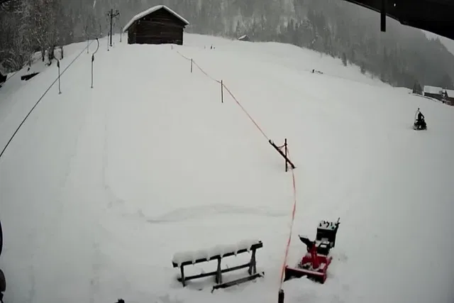 Winterszene mit Holzhütte auf verschneitem Hang, Absperrseile, Bank und Skiausrüstung im Vordergrund
