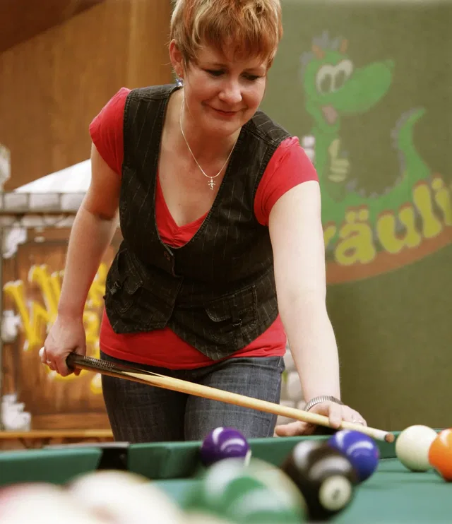 Frau mit rotem Shirt und dunkler Weste spielt Billard, konzentriert beim Stoß.