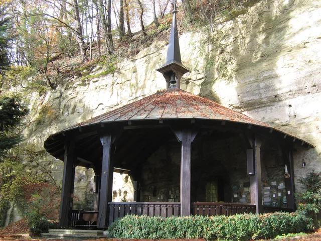 Runder Holzpavillon unter Felswand, Spitzdach, Turmspitze, umgeben von Bäumen