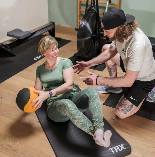 Frau sitzt auf Matte mit Medizinball, Trainer gibt Anleitung im Fitnessstudio.