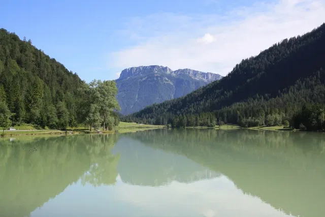 Ruhevoller See im Tal, von Wald umgeben, mit Bergen im Hintergrund und spiegelndem Wasser.