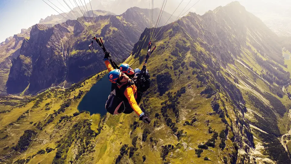 Pilot und Passagier beim Tandem-Gleitschirmflug vor felsigen Alpengipfeln.