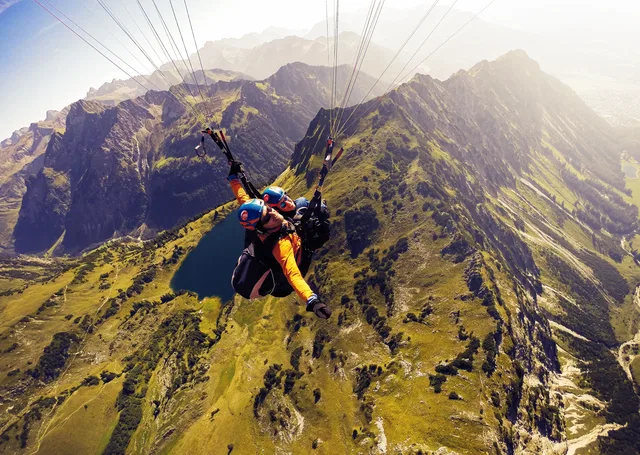 Pilot und Passagier beim Tandem-Gleitschirmflug vor felsigen Alpengipfeln.