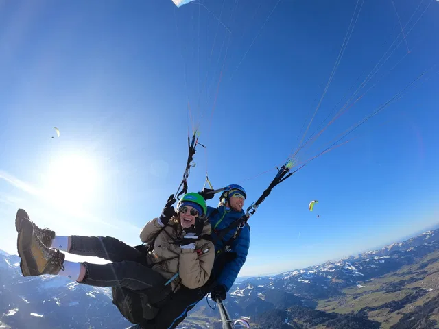 Passagier und Tandempilot über einer Berglandschaft unter blauem Himmel