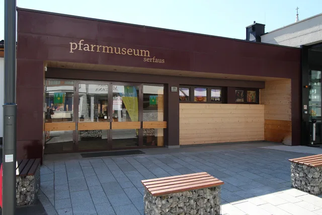 Außenansicht des Pfarrmuseums Serfaus mit Glasfront, Holzverkleidung und Sitzbänken im Vorhof.