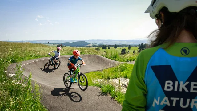 Zwei Kinder fahren auf einem Pumptrack durch eine grüne Landschaft, Betreuerin beobachtet von hinten