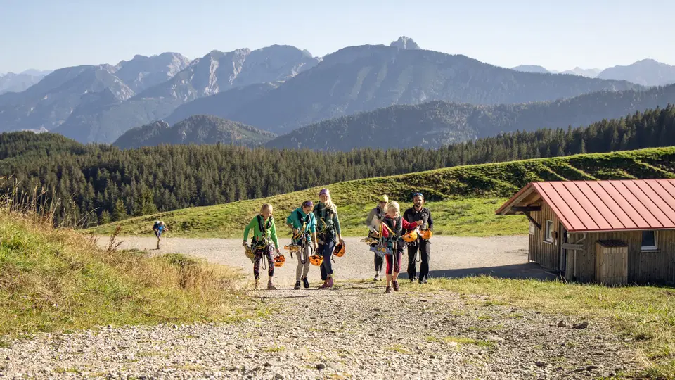 Gruppe Wanderer mit Rucksack geht einen Kiesweg entlang vor einer Berglandschaft und einer Hütte.