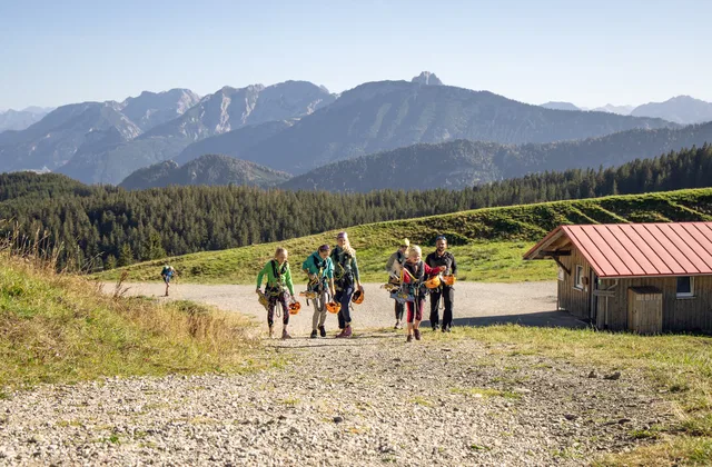 Gruppe Wanderer mit Rucksack geht einen Kiesweg entlang vor einer Berglandschaft und einer Hütte.