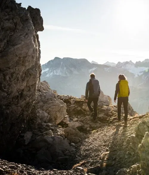 Zwei Wanderer mit Rucksäcken auf felsigem Gebirgspfad vor Bergkulisse.