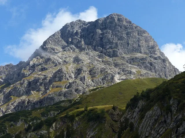 Felsiger Berggipfel unter blauem Himmel mit grasbewachsenen Hängen.