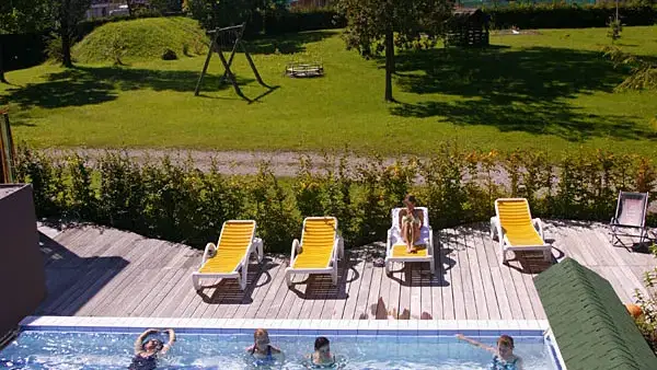 Außenpool mit mehreren Schwimmern, Liegen am Beckenrand und grünem Garten mit Bergen dahinter.