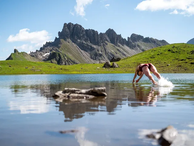 Person steigt ins Wasser eines Bergsees; grüne Hänge und schroffe Bergkämme im Hintergrund.