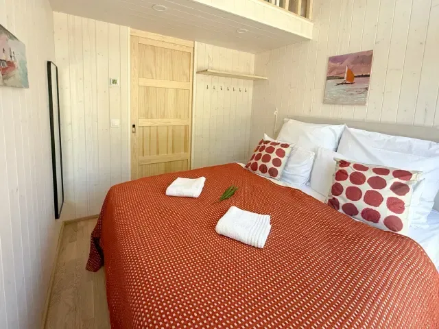 Doppelbett in weißem Holzraum mit oranger Bettdecke, weißen Kissen mit roten Tupfen und zwei Handtü-