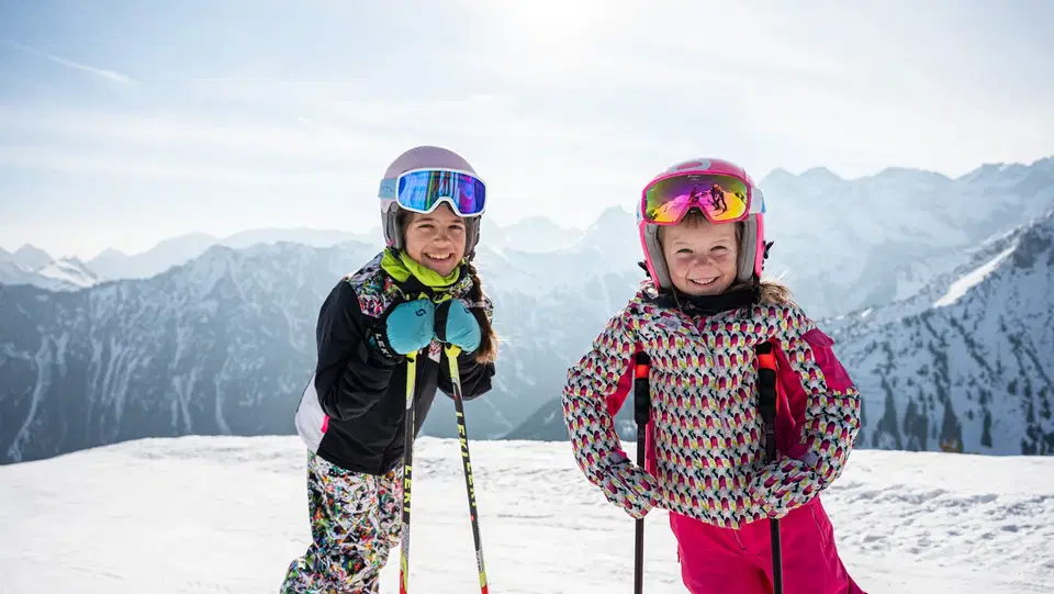 Zwei Kinder in Skikleidung mit Helmen posieren auf verschneiter Bergpiste.
