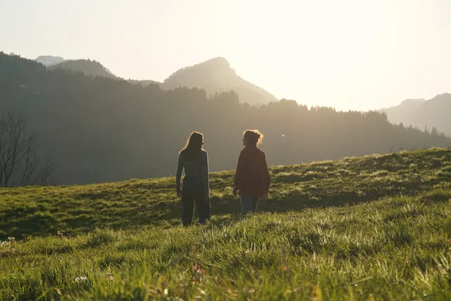 Zwei Menschen auf einer Wiese vor bewaldeten Bergen im Gegenlicht