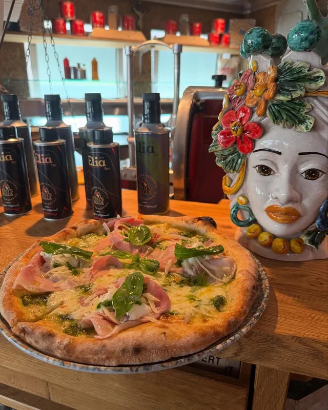 Pizza mit Schinken und Rucola auf Holztisch in einer Bar, daneben eine bemalte Keramikfigur.