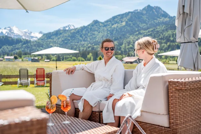 Zwei Frauen in Bademänteln sitzen entspannt im Outdoor-Loungebereich vor Berglandschaft.