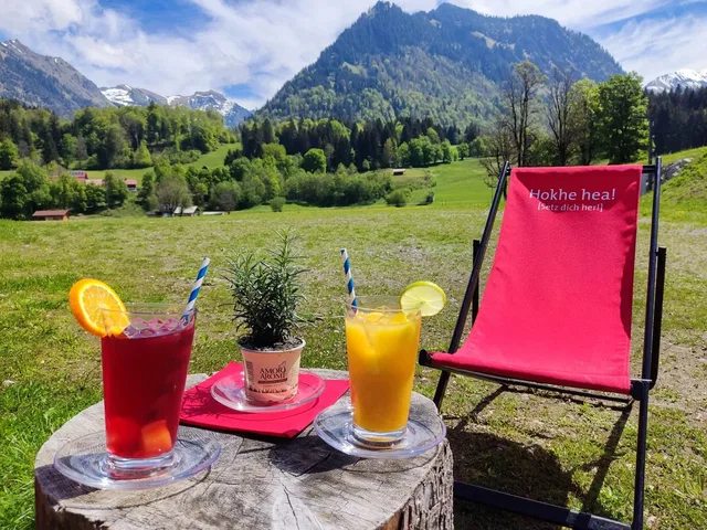 Bunte Getränke auf Baumstumpf-Tisch mit Pflanze und rotem Liegestuhl vor Berglandschaft.